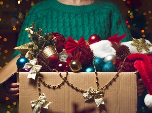 A person wearing a green sweater holding a large box full of holiday decorations.