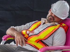 A worker wearing a safety vest and hard hat falling asleep in a chair at work.