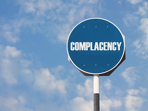 A blue sign that says "Complacency" against a blue sky with clouds.