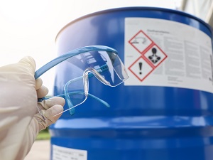 A gloved hand holding safety glasses near a 55-gallon drum with hazardous chemicals.