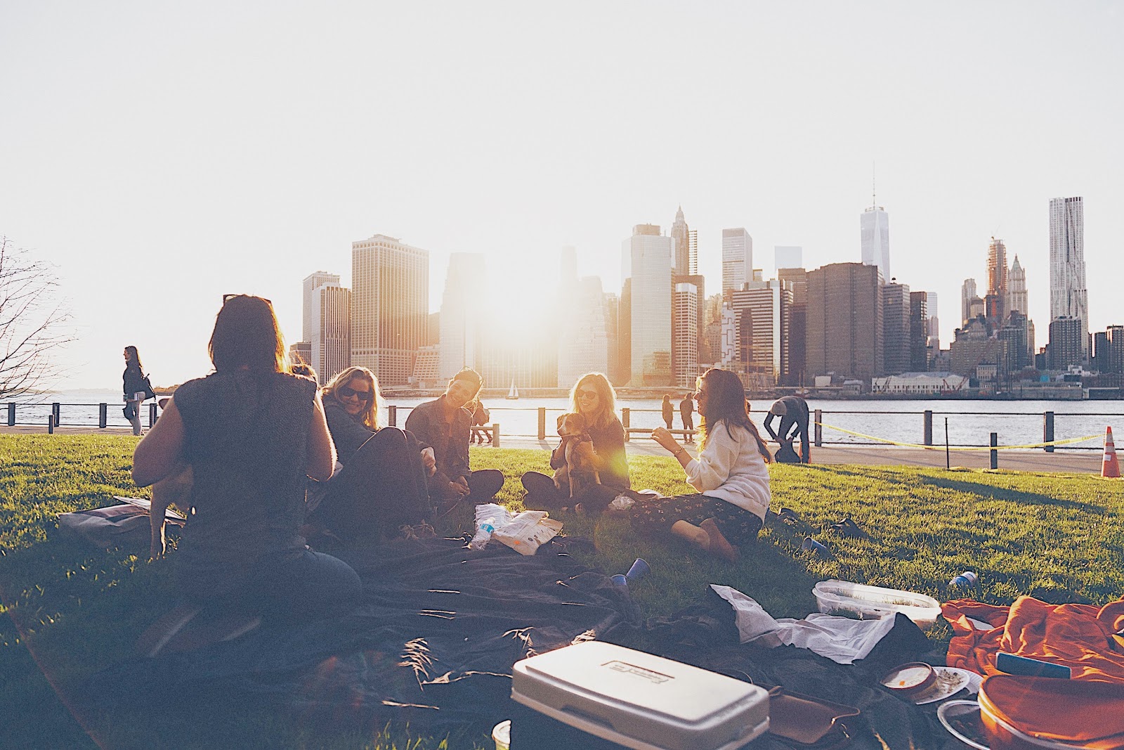 A group of 6 friends share a sunset picnic together in a grassy park across the river from a large city with sunset shining through a skyline filled with tall high rise buildings
