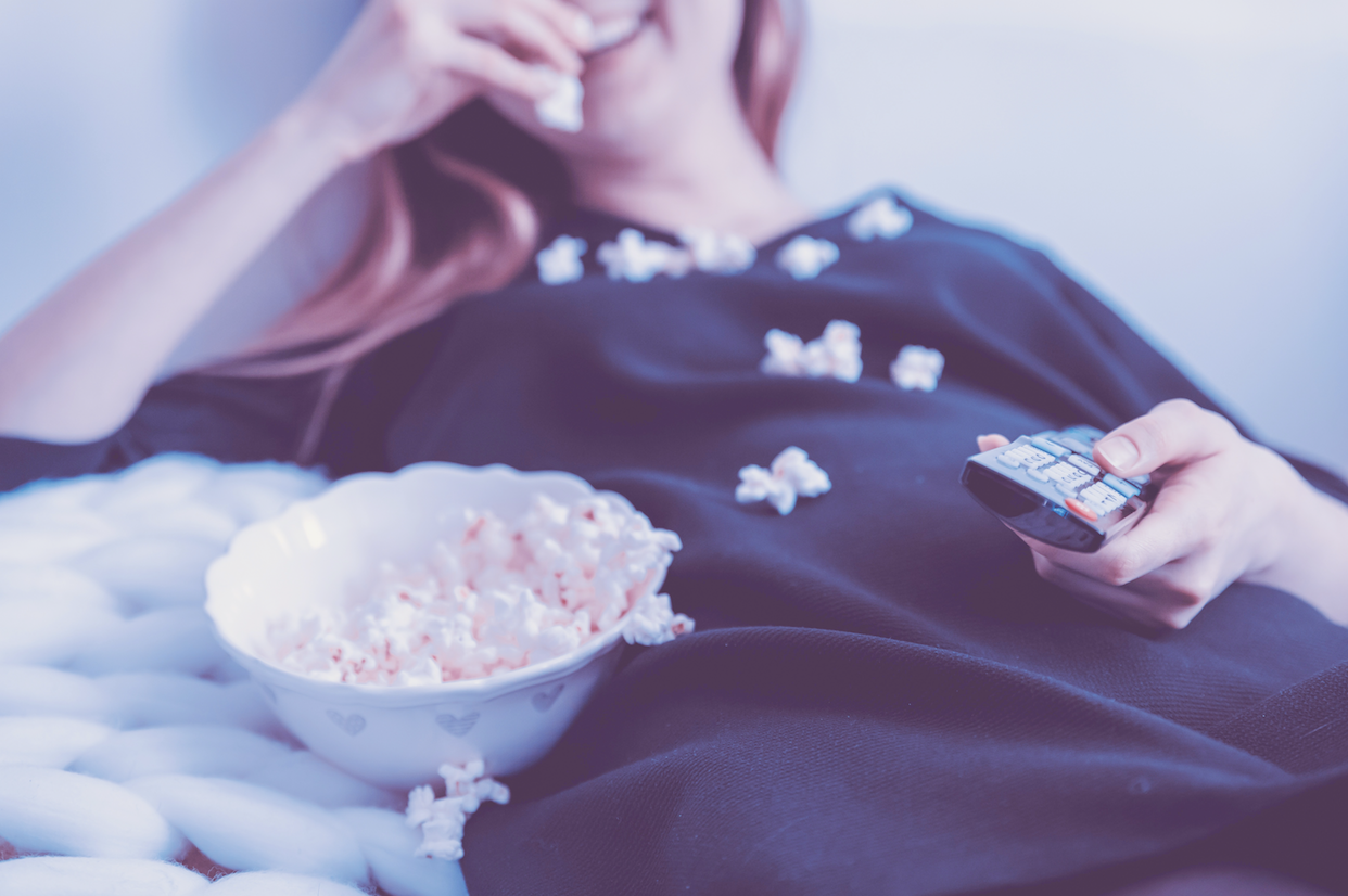 female lounges happily with tv remote in hand beside a bowl filled with popcorn as she snacks and watches a favorite movie