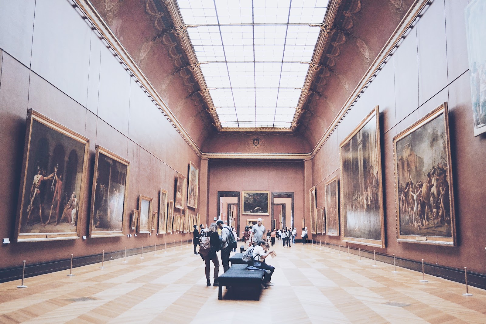 people stand and sit gathered on benches within a long museum hallway with tall walls lined with large artwork