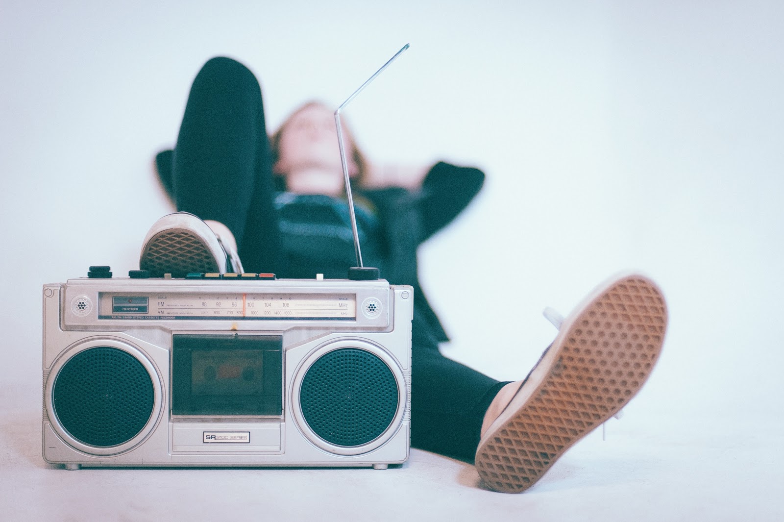 a young female lounges on a bed casually with one foot resting on top of a silver AM/FM radio playing her favorite music