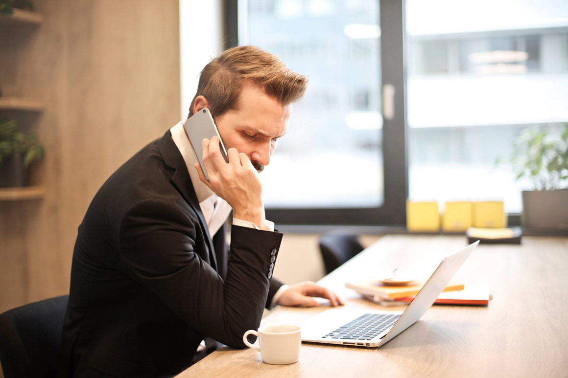 A man wearing office attire talking on his phone in front of his computer.