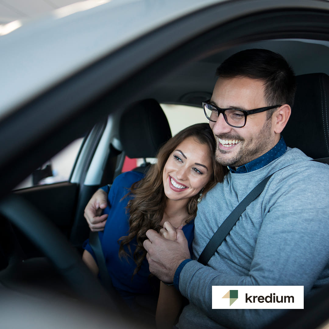 Momak sa naočarima i devojka nasmejani i zagrljeni sede u automobilu držeći se za ruke.