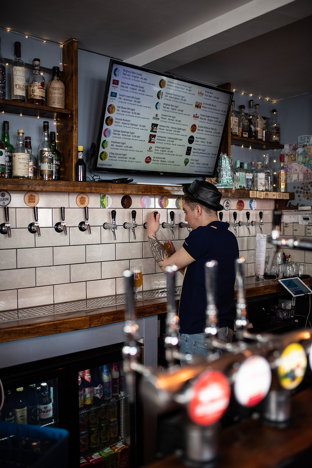 The bar at Brighton Beer Haus has a wall of keg taps set into white metro tiles. There is a large television screen which shows the wide array of beers available on the menu. The bar person is pouring a beer from one of the keg taps.