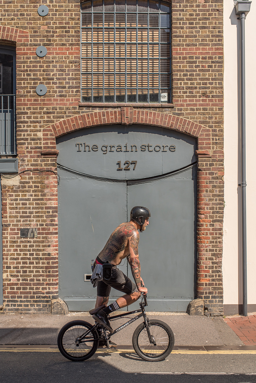 A topless tattooed man in shorts rides in front of The Grain Store on a BMX