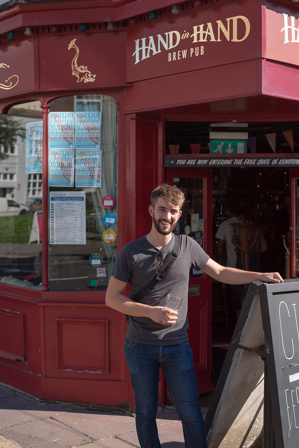 Owner of the Hand in Hand pub in Brighton poses outside of the pub with a pint in one hand. He has his other hand resting on top of a A board.