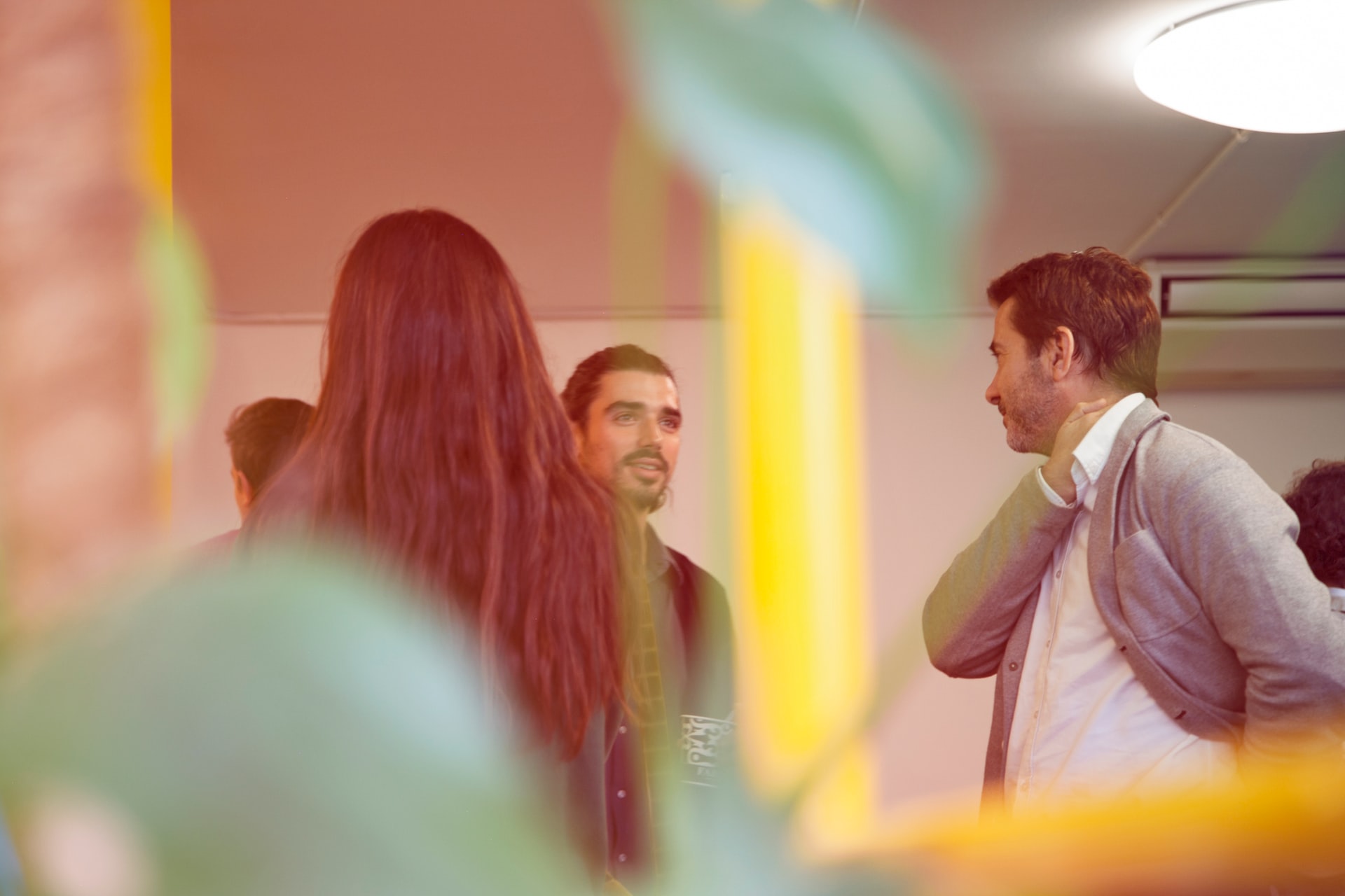 foreground is an extremely blurry green plant. In between stems are people in business casual dress. A man in the middle smiles at a man on the right of frame. A blurry yellow pillar separates them.