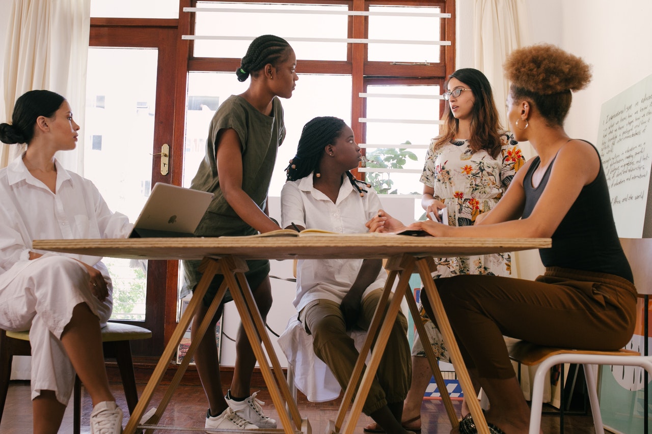 Team of dark, medium, and light-skinned women stand around a table having a discussion about hiring for diversity.