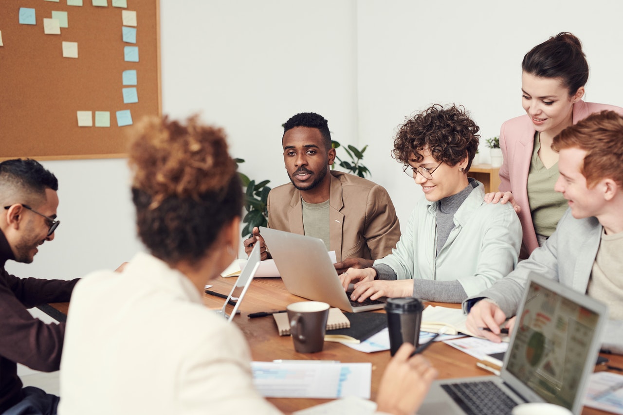 Team of dark, light, and medium-skinned people dressed in business casual sit around a table discussing hiring for diversity.