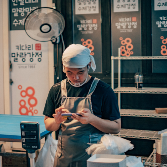 A factory worker wearing a hair net holds a cell phone in his hand to check his work on the factory floor.