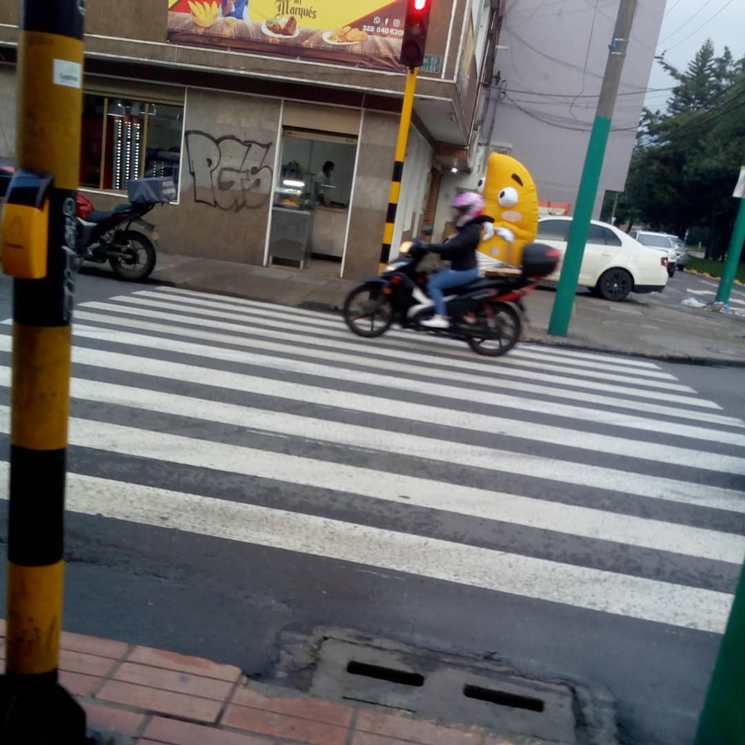 Al otro extremo de la calle una motociclista pasa al pie de una persona con disfraz de empanada.