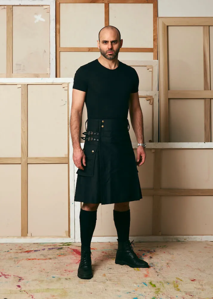 Portrait of Alias Trate, wearing black and standing in front of painting canvases.