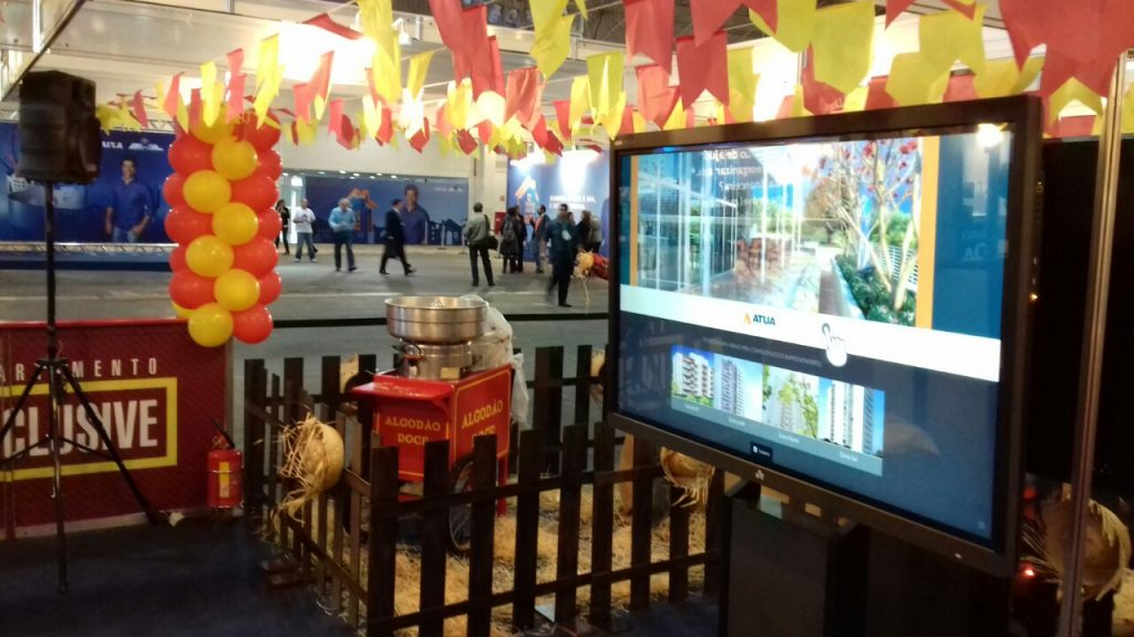 Estande da Construtora Atua no Feirão da Caixa, com pipoca, bandeirolas de festa junina e Showcase Imóveis, o catálogo interativo da Aqua.