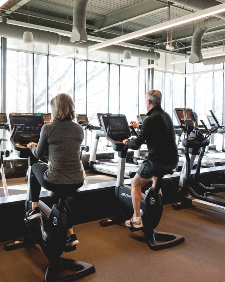 Medium shot of a middle-aged couple exercising on stationary bikes in a gym.