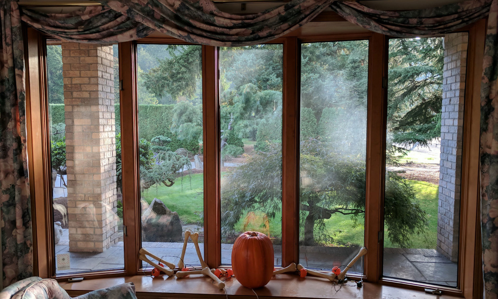 A bay & bow window with clean, clear glass and brown, stained trim overlooks a yard of greenery. There are Halloween decorations and a big, orange pumpkin sitting interiorly in front of the window.