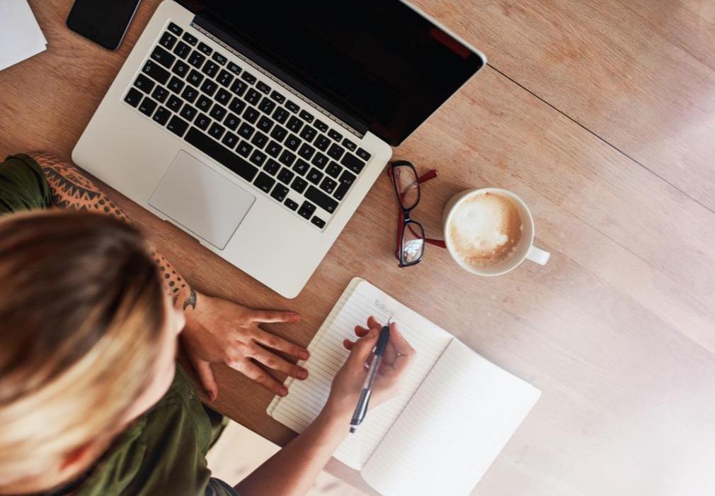 A woman sits in front of her laptop writing notes about tasks that need to be completed.