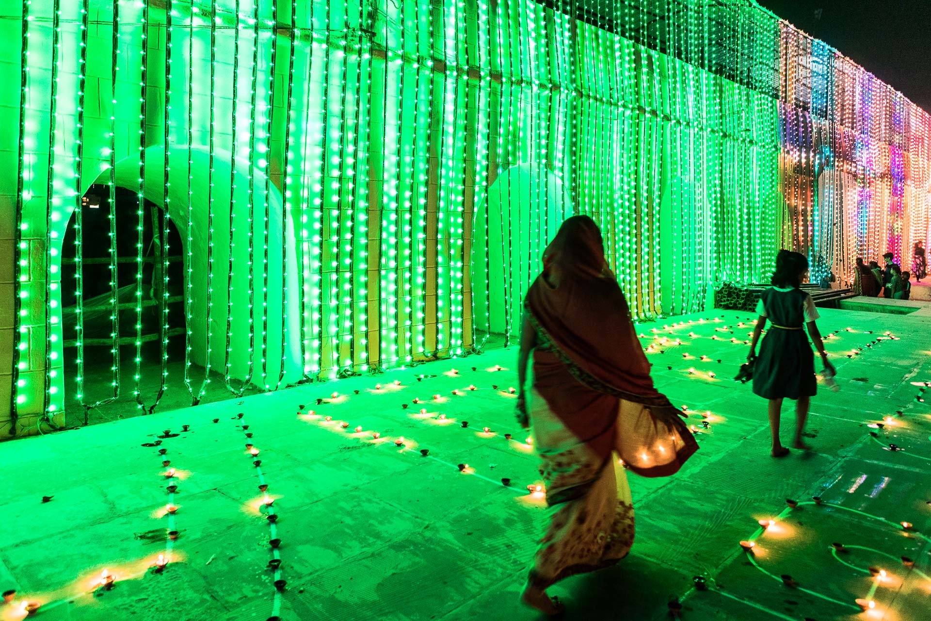 Two people walk past candles on the ground with a heavy green light source in the background