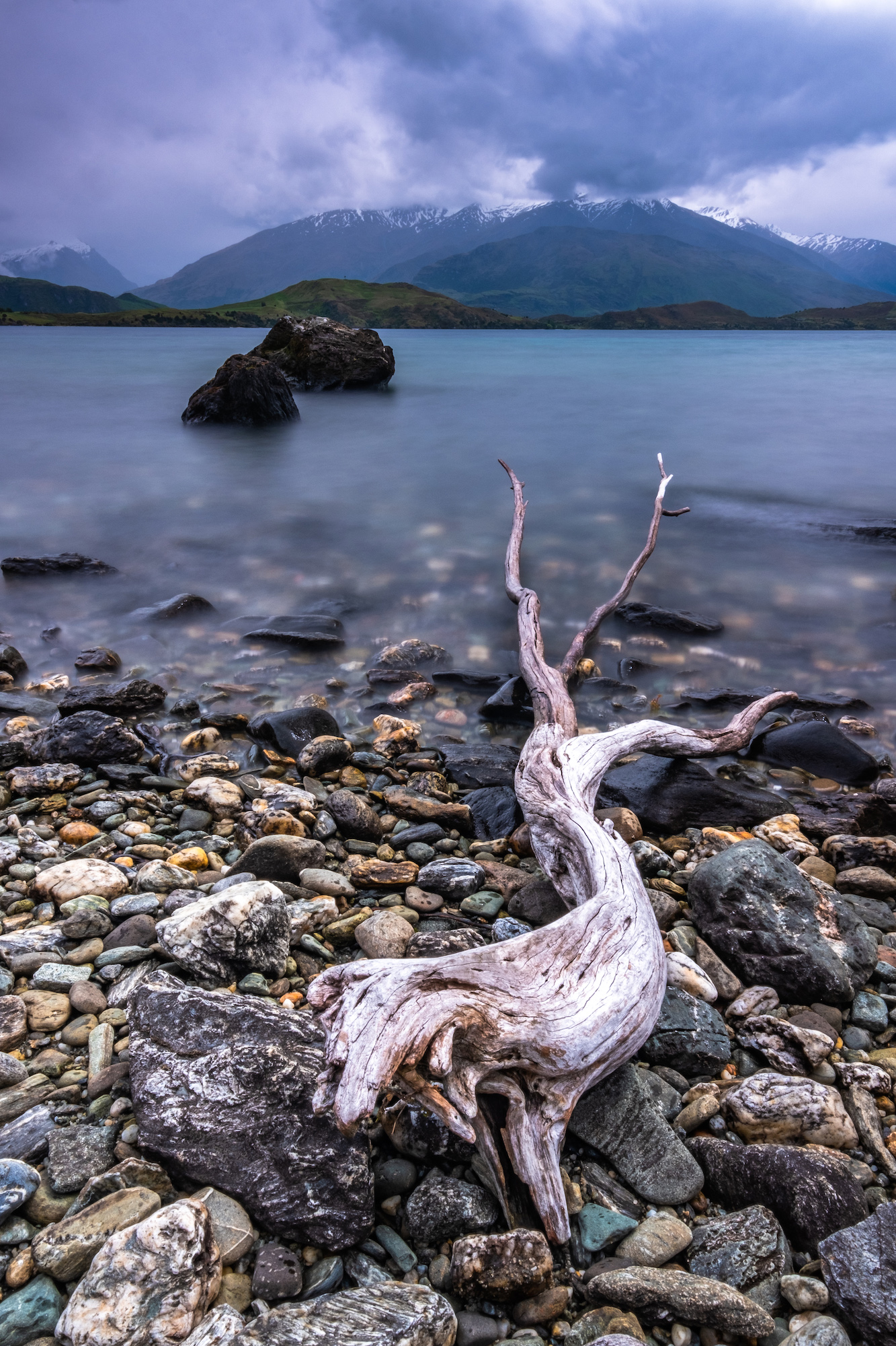 Washed up driftwood and pebbles make for a textured foreground as The Remakables are almost swallowed by low clouds; Queenstown, New Zealand