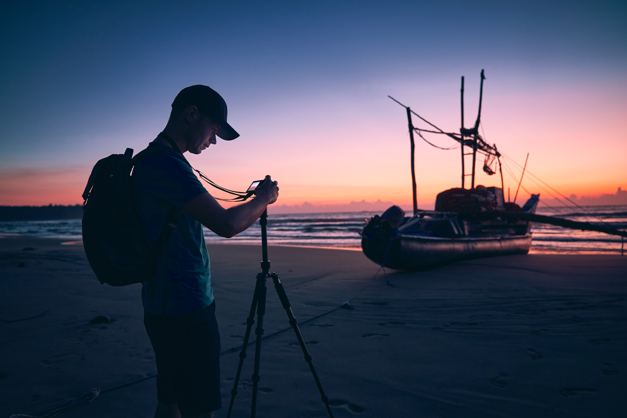 Silhouette of young photographer (traveler) with tripod on beach against fishing boat and sea. Beautiful sunrise in Sri Lanka.