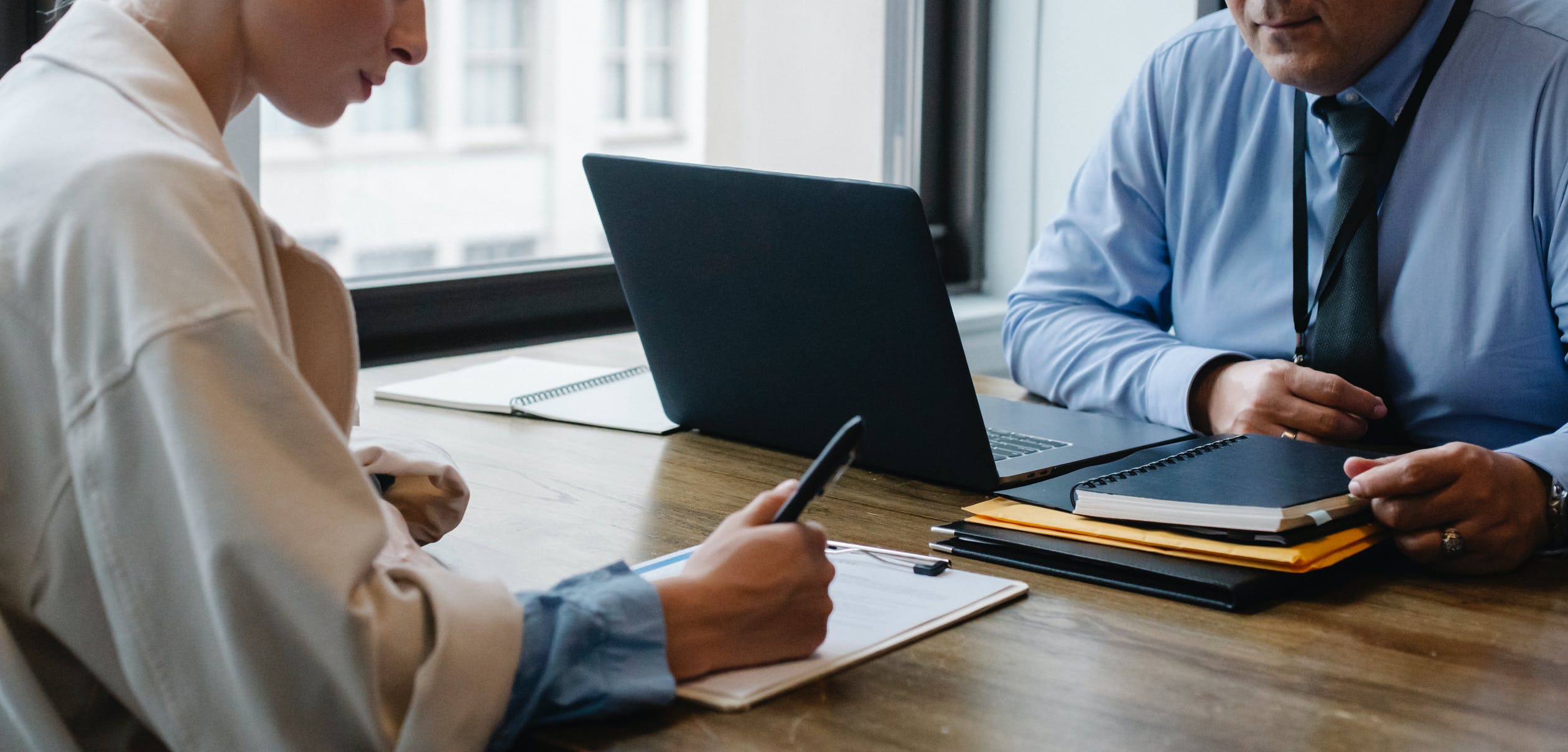 A business woman completing a clipboard form on a wooden table across from a businessman seated in front of a laptop and folders.