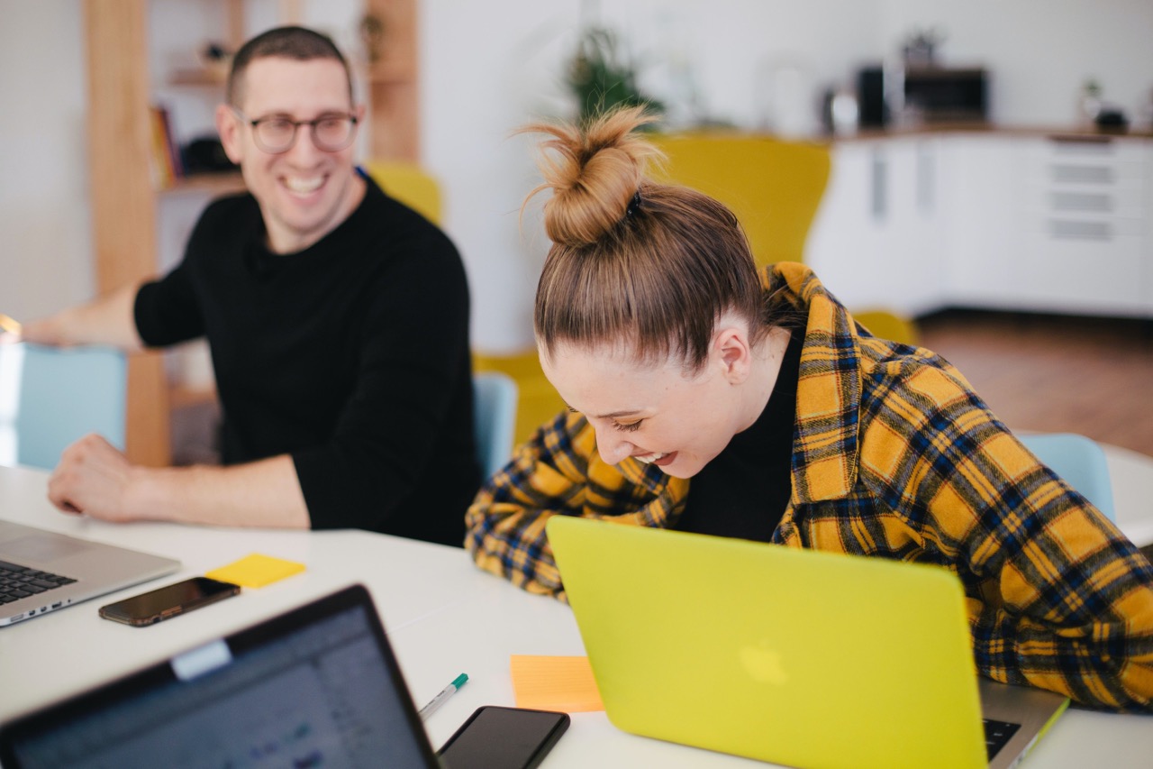 Woman in yellow check jacket laughing with her male colleague seated in front of their laptops.