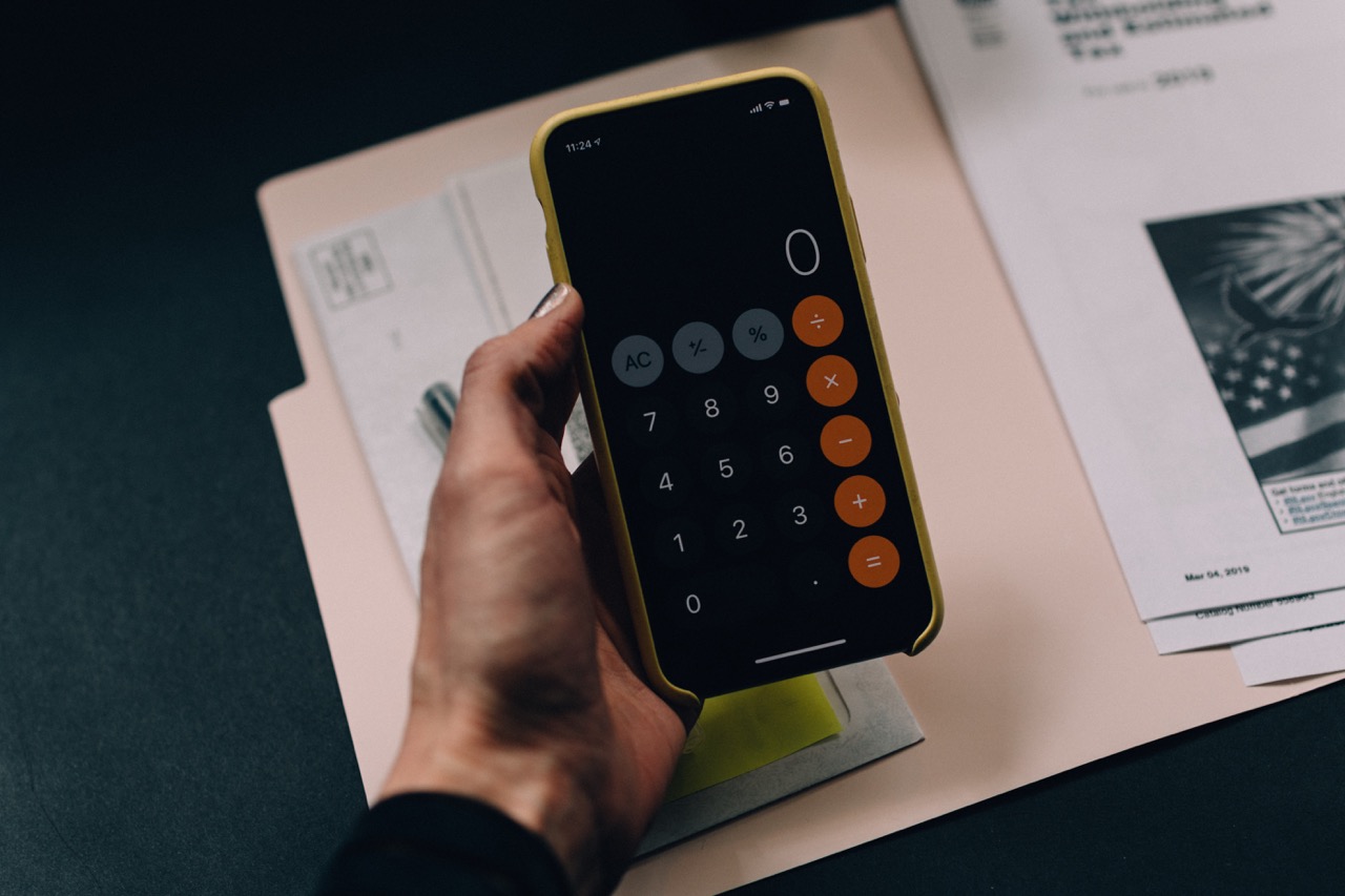 Hand holding a mobile phone displaying a calculator in front of a file containing bills and paperwork.