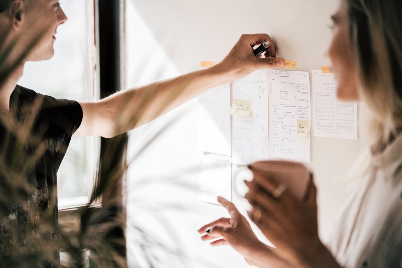 A woman holding a cup looking at a man pinning papers to a wall.
