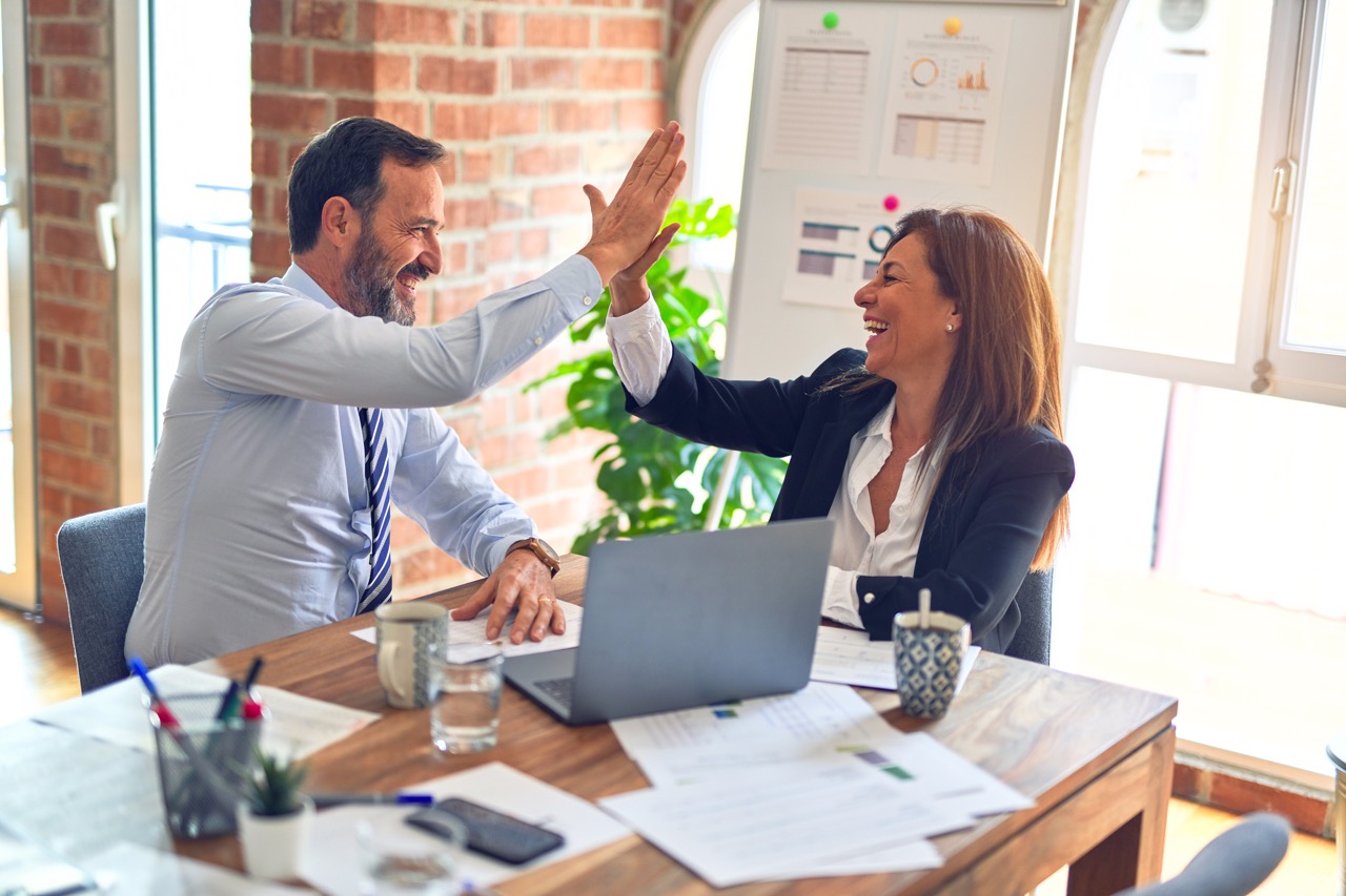 Man and women in a bright office building high fiving at a desk with papers and a laptop.