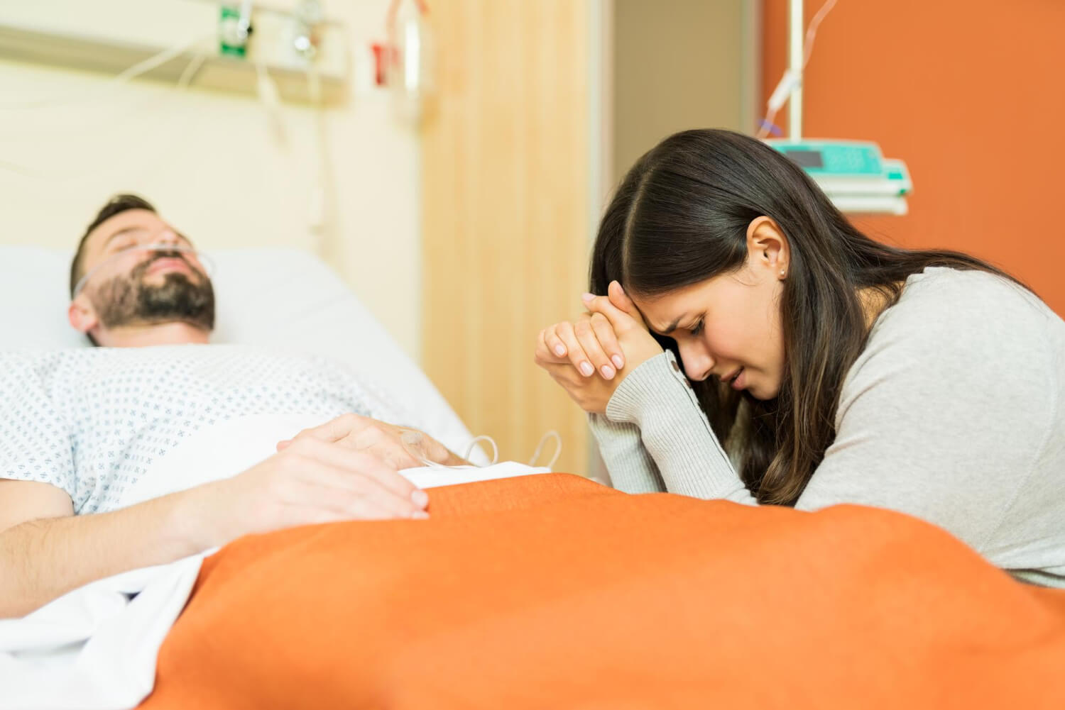 Crying woman praying for male patient while sitting by bed at hospital.