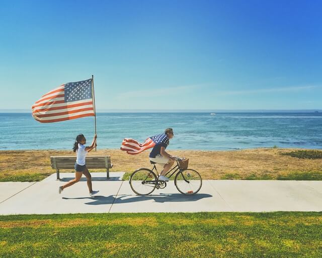 Hombre montando en bici por una acera con una bandera de EE.UU. como capa. 