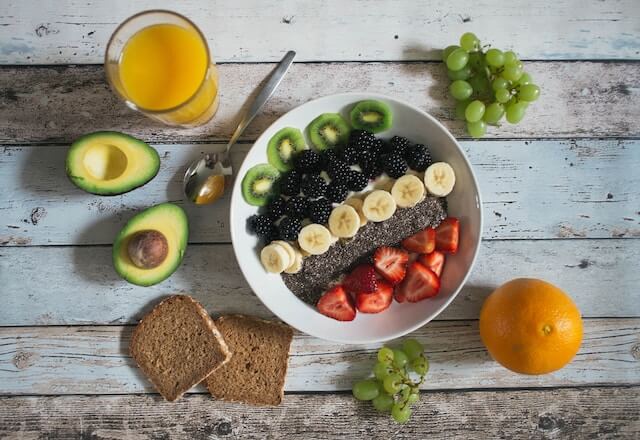 Wooden pale table with a bowl of several fruits, avocado, orange juice and wheat bread. 