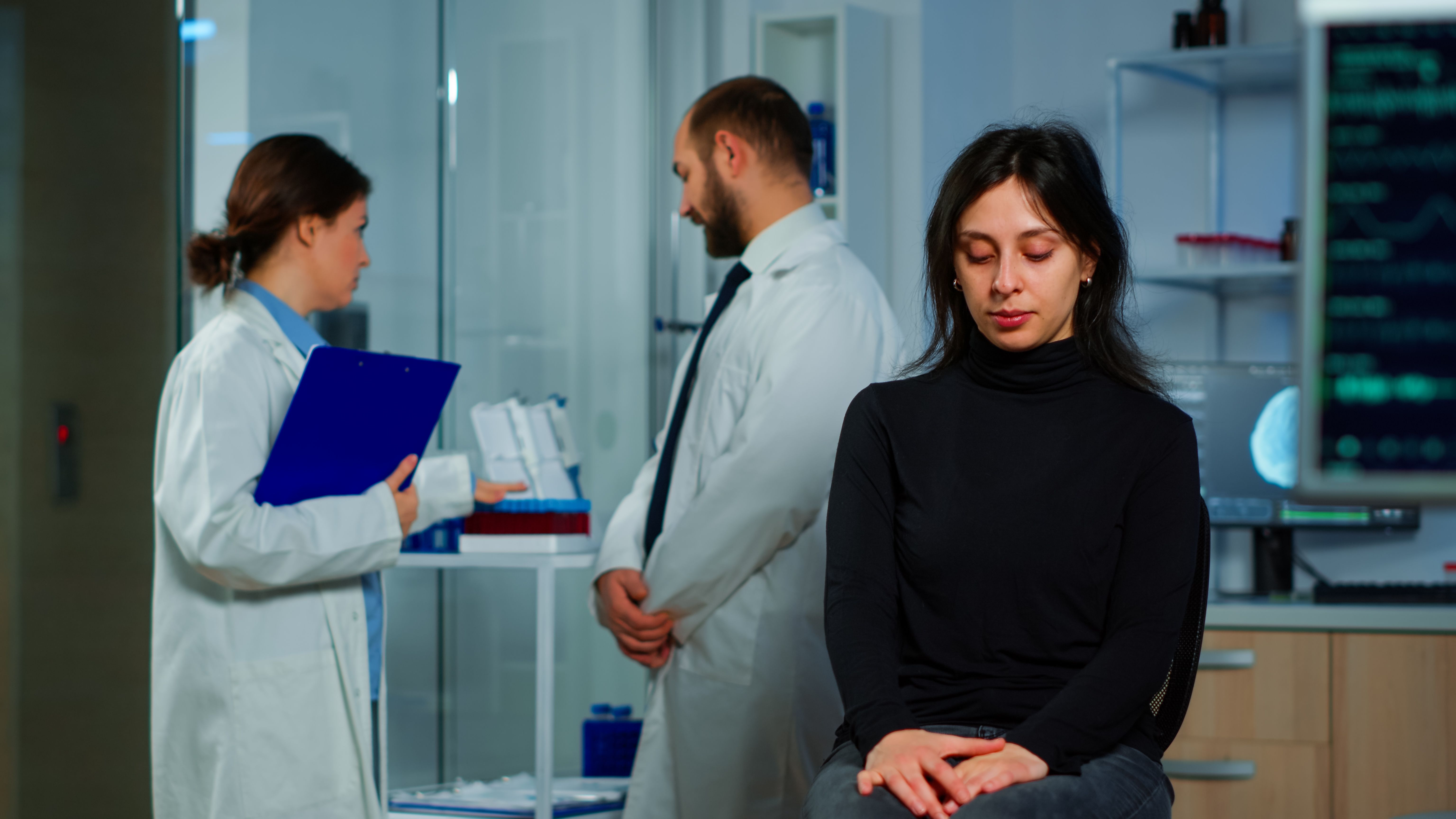 Negligencia médica mujer preocupada con dos médicos hablando detrás de ella esperando resultados 