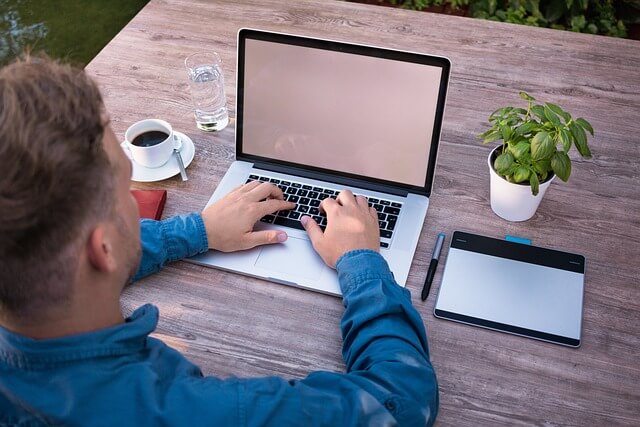 Man sitting on a table with a laptop, notebook, plant and coffee. 