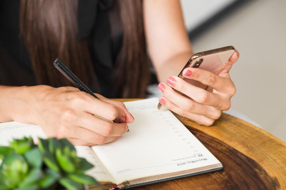 Mujer tomando notas en su agenda utilizando su teléfono. 