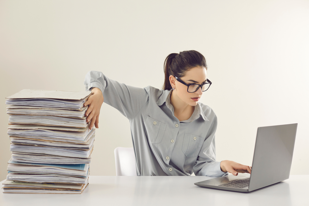Young accountant working on laptop computer sitting at desk with pile of papers. Paperwork vs electronic documents.