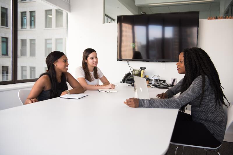 alt text: Um equipe de mulheres está em reunião sentadas em volta de uma mesa branca.