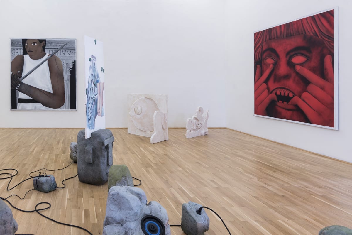 the image shows a room in the gallery with large paintings one of a woman and one of a scary red head. On the floor there is an installation of historic looking heads.