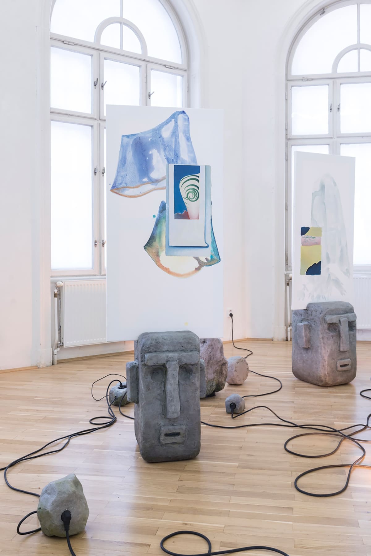 Image shows a gallery room with installations on the floor, cables, paintings on material hanging from the ceiling
