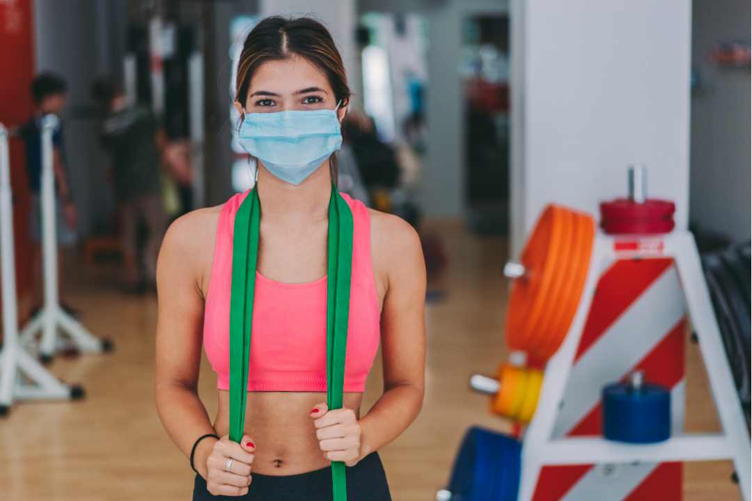 A fit lady wearing a mask and standing in a gym with a resistance band around her neck