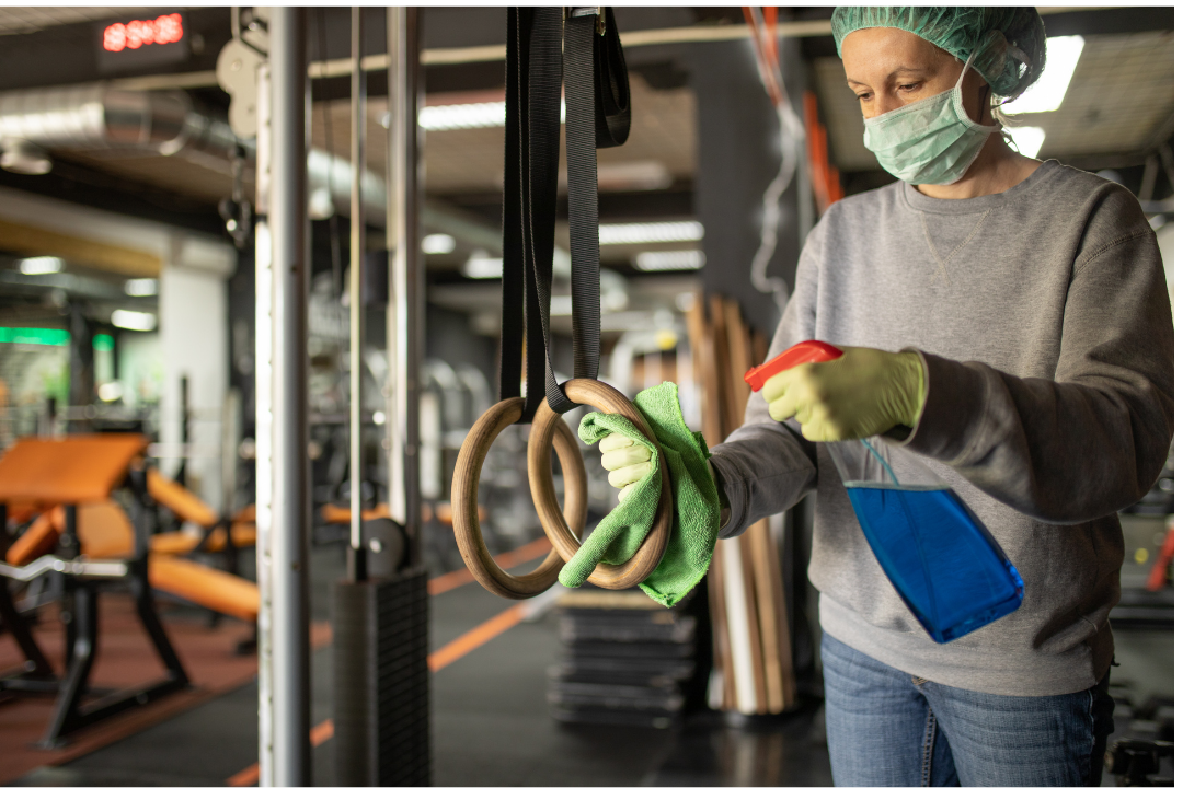 Woman wearing gloves and safety mask, sanitizing gym equipment using disinfectant
