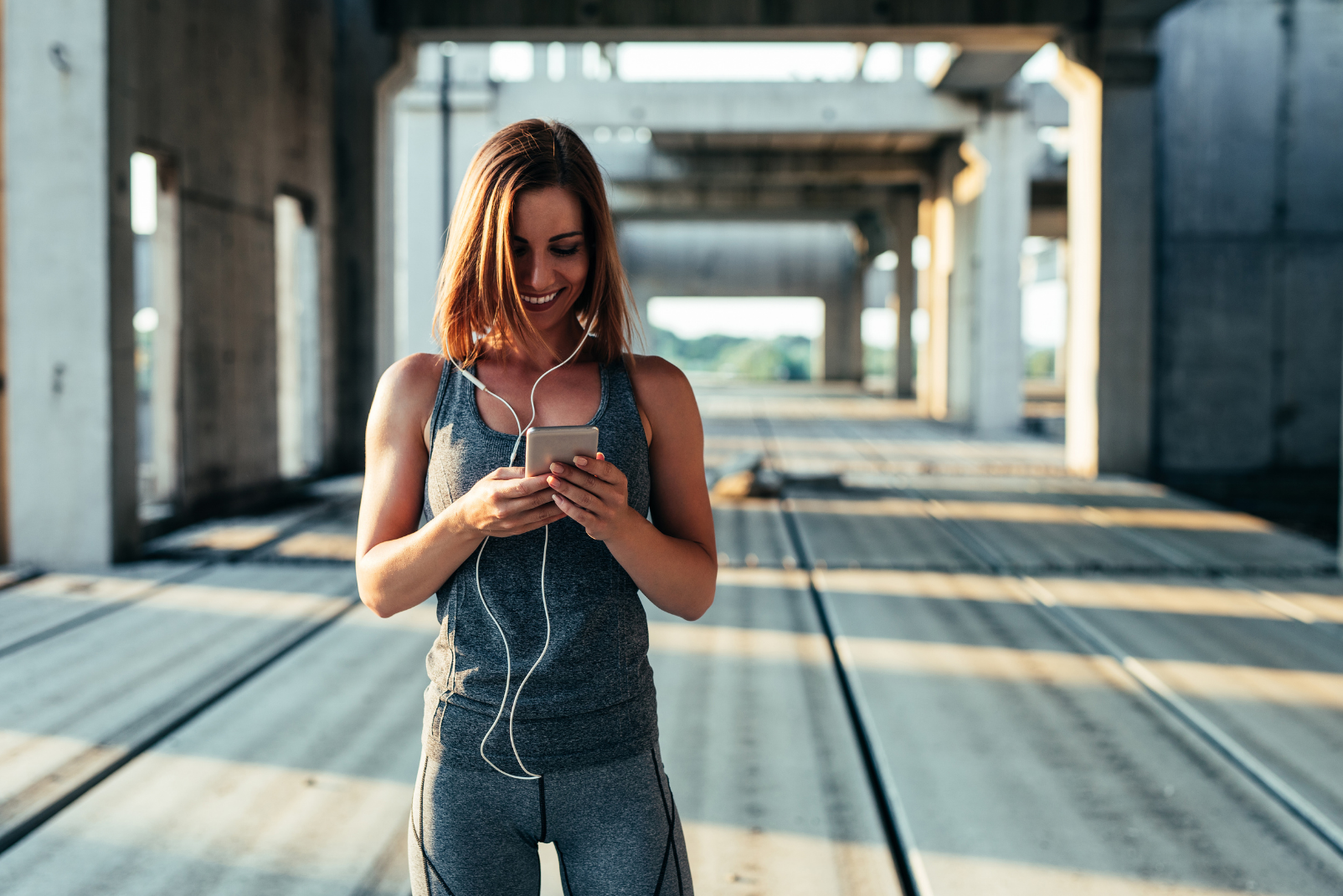 A happy woman in gym clothing, smiling and looking at her phone while standing on a wide bridge