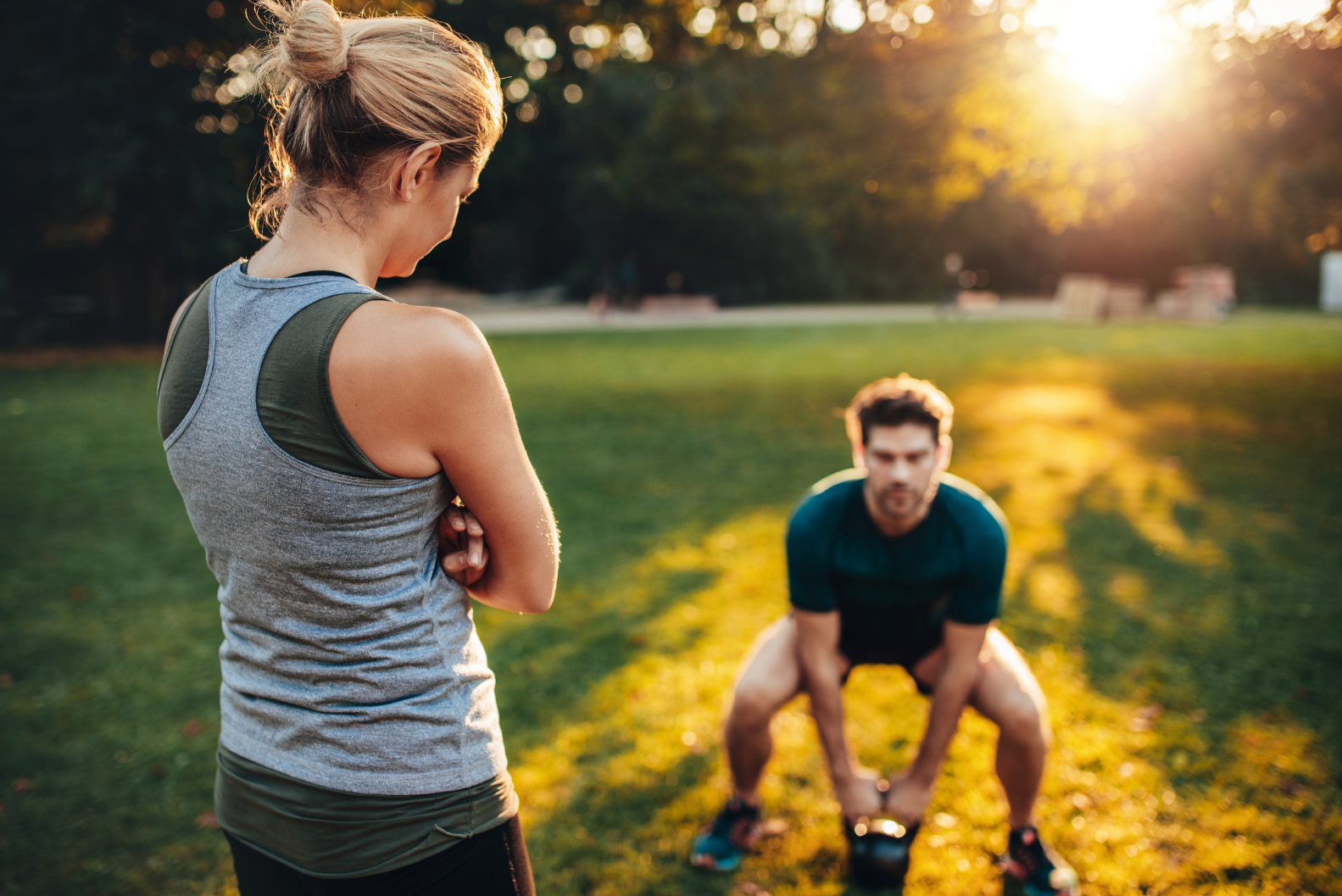 A female fitness trainer overseeing a male student as he swings a kettle-ball in an open park