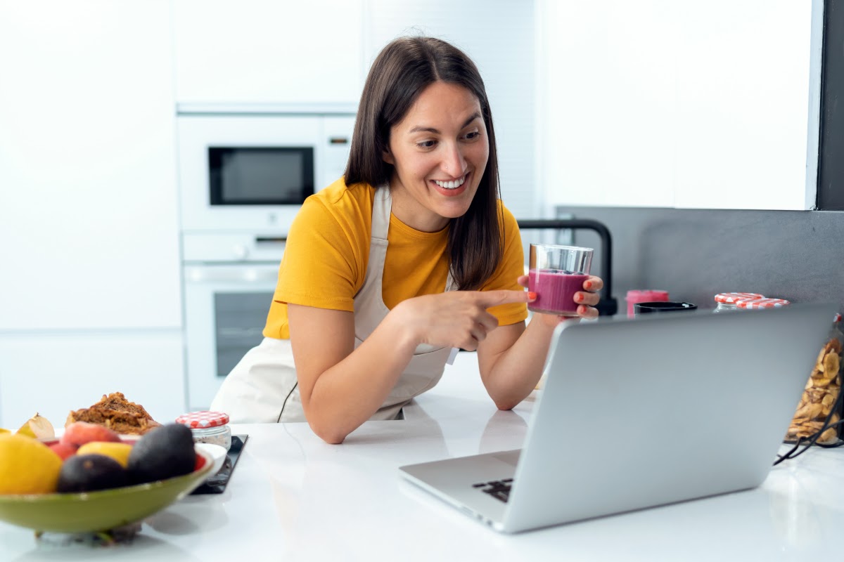 Smiling woman holding a glass of fruit juice and pointing at it while looking at her laptop