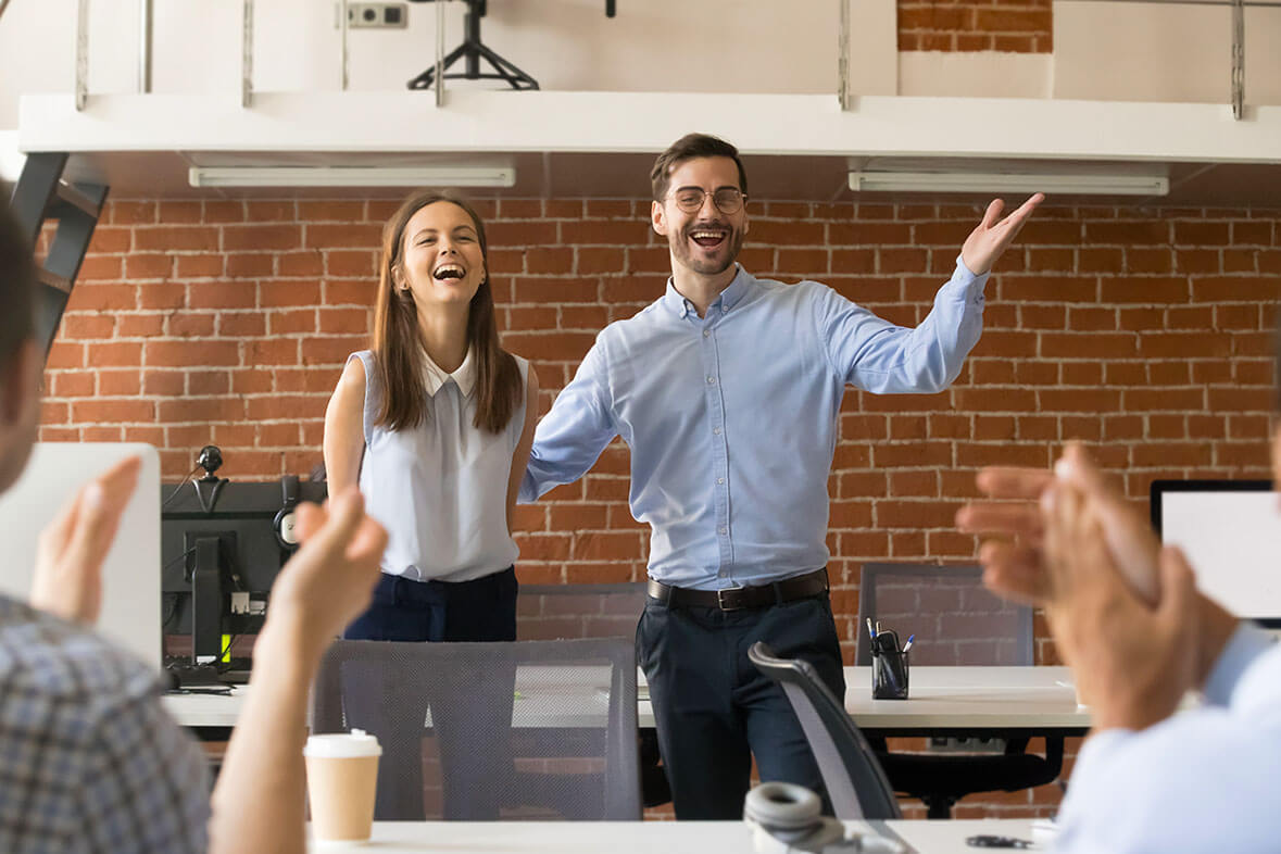 Smiling man recognizes woman in front of applauding office.