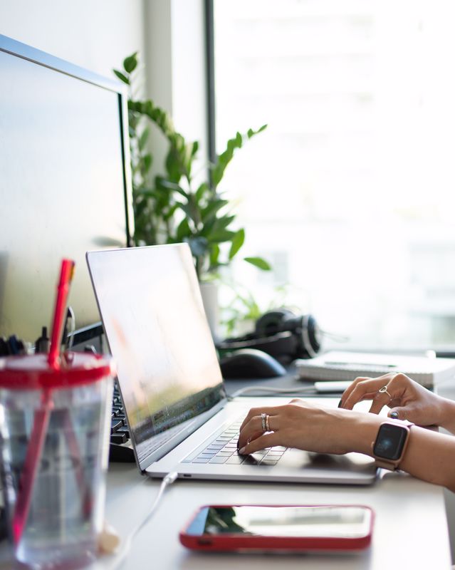 Mains d'une personne en télétravail qui écrit sur un clavier d'ordinateur portable posé sur un bureau avec un téléphone, un gobelet en plastique, un deuxième écran et une plante située en fond