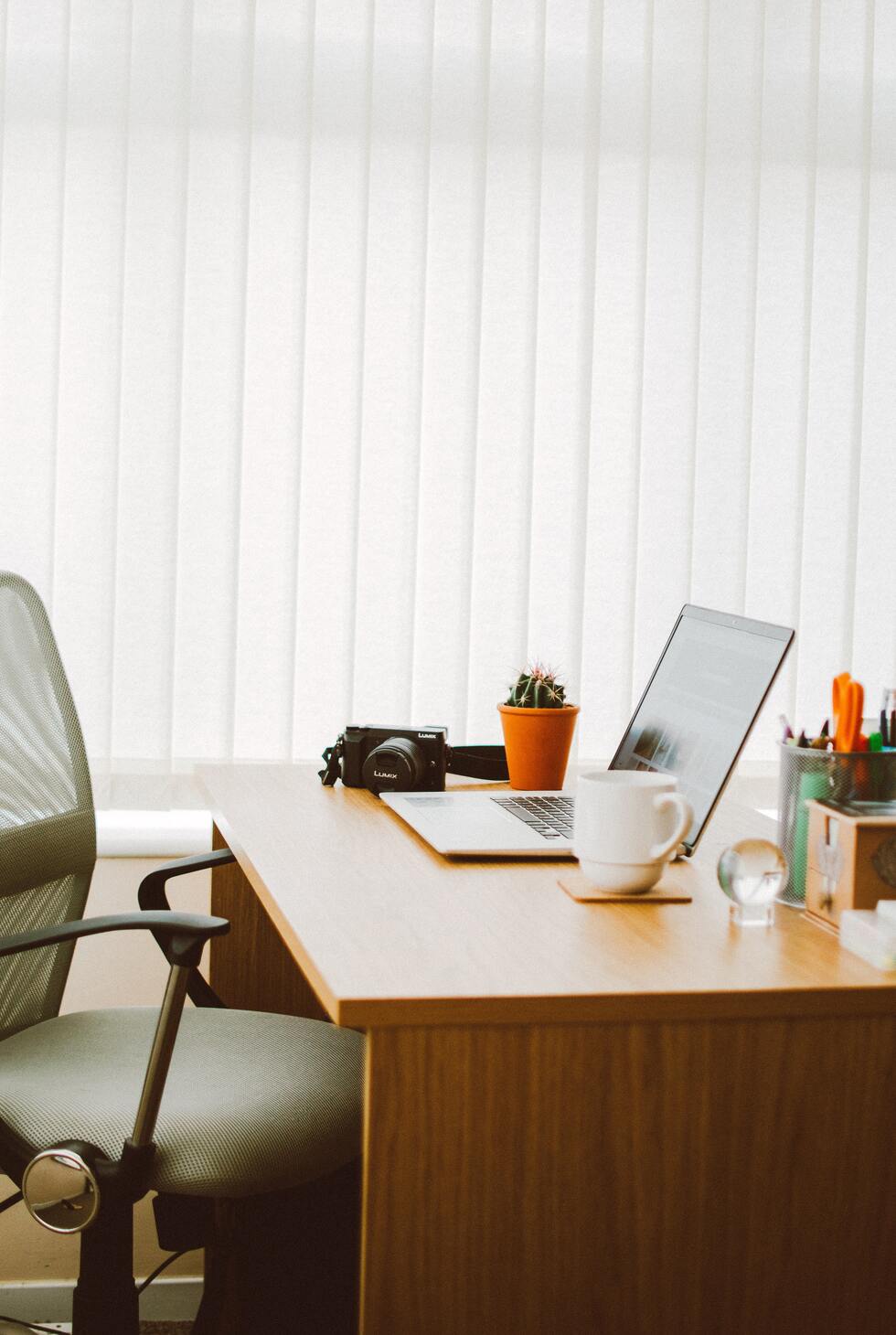 Un bureau en bois avec un ordinateur, une tasse, une plante, un appareil photo, des stylos et des ciseaux.