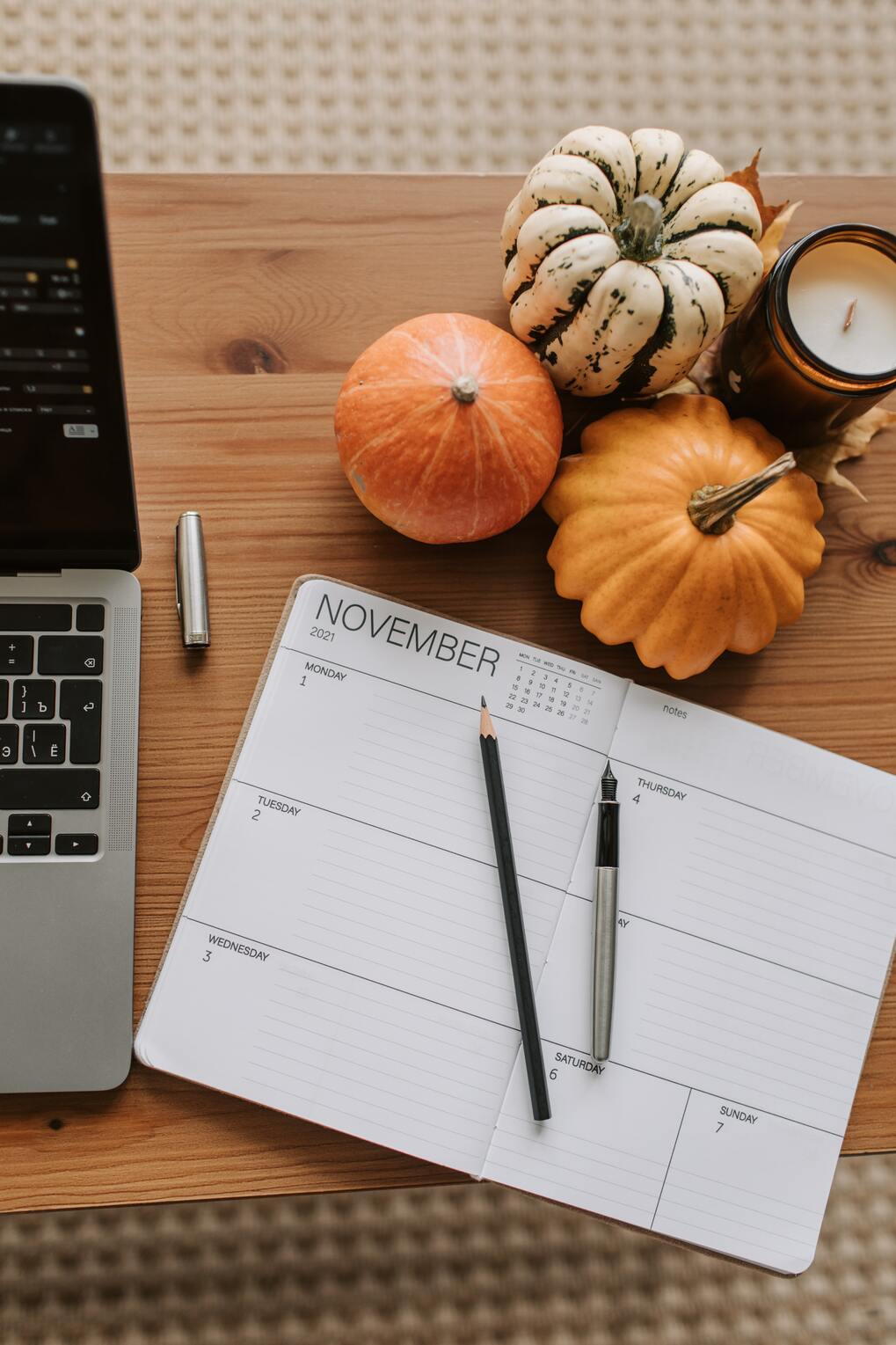 Un table en bois avec un calendrier papier ouvert au mois de novembre, un ordinateur, une bougie et trois citrouilles de différentes couleurs.